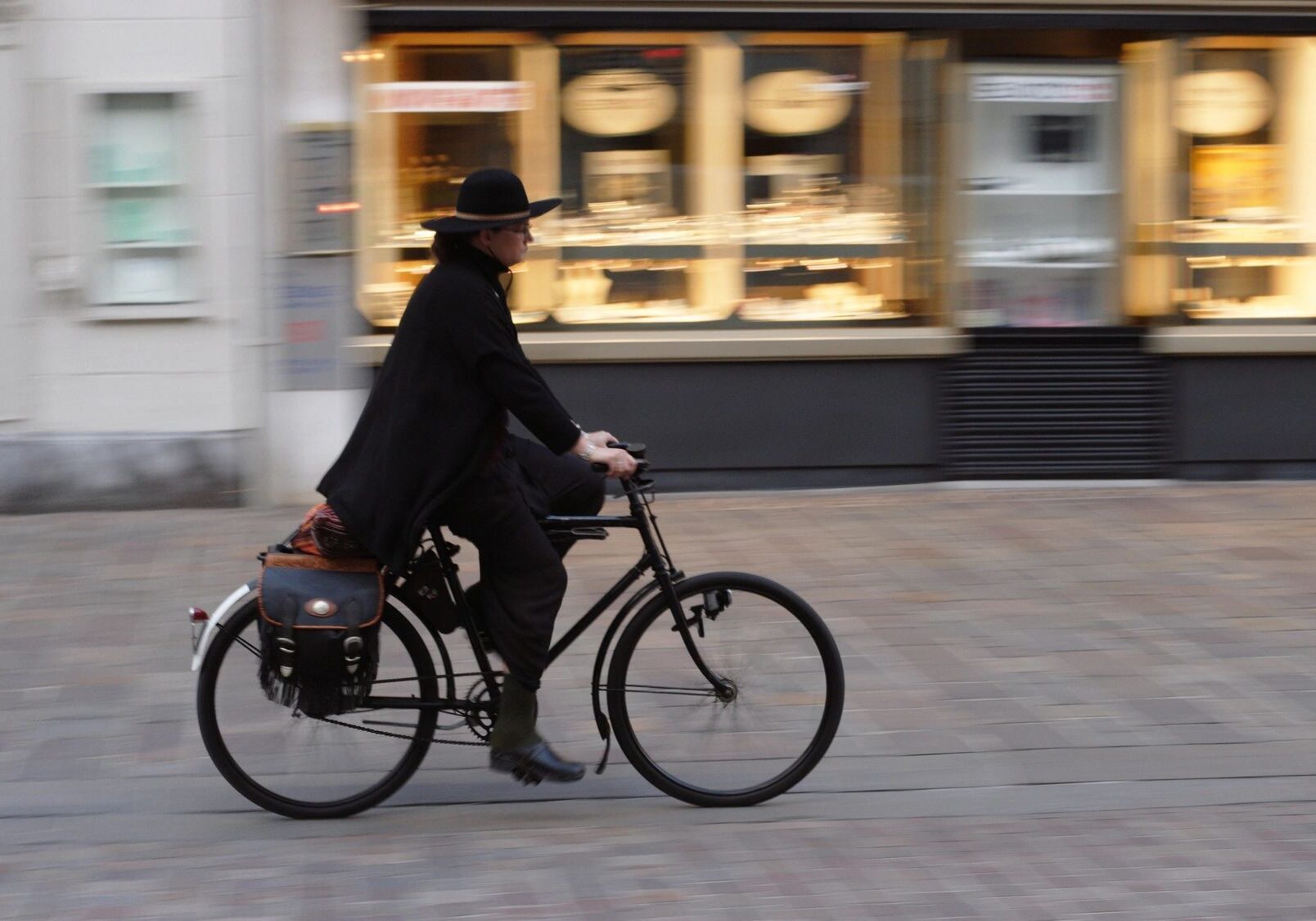 Person cycling past blurred storefronts, wearing hat.